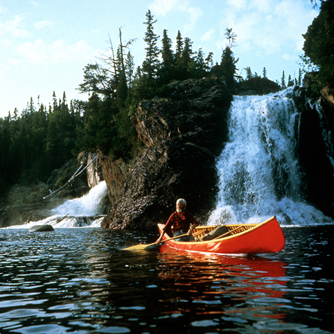 Colour photograph of a man paddling in a red canoe on a lake, with a rocky cliff and waterfall behind him.