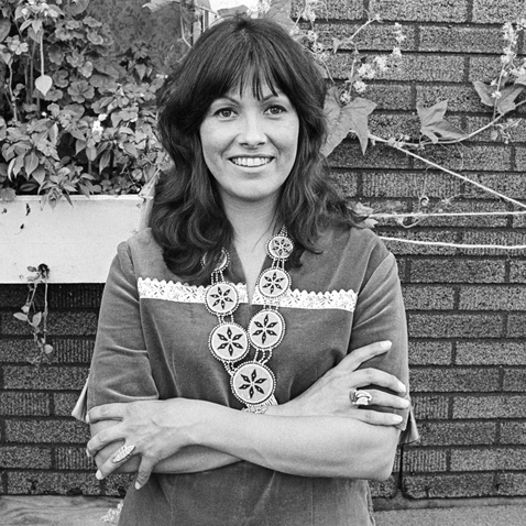 Black and White photograph of a woman with arms crossed standing in front of a brick building with vines and foliage attached.