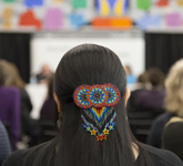 Colour photograph of a multi-coloured, beaded hair clip decorating the back of a woman’s head. The woman is sitting in the Pellan Room of Library and Archives Canada, listening to a panel of speakers.