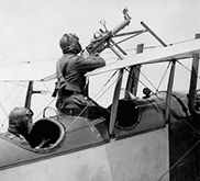 Black and white photograph showing a biplane, with the pilot aiming a machine gun and an observer sitting in the rear.