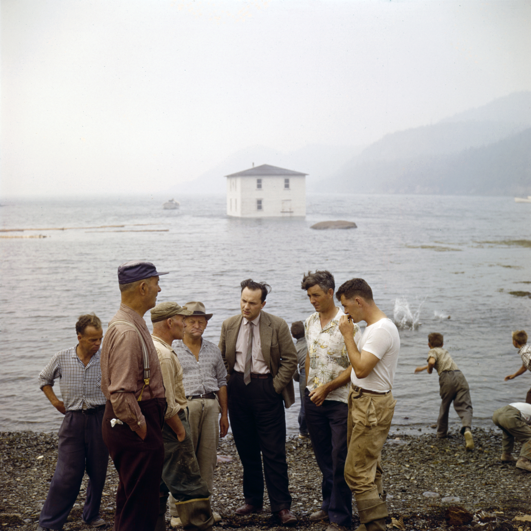 Several men talking on a beach. To the right of the image, children are throwing rocks into the water. In the background, a house is floating by in the water.