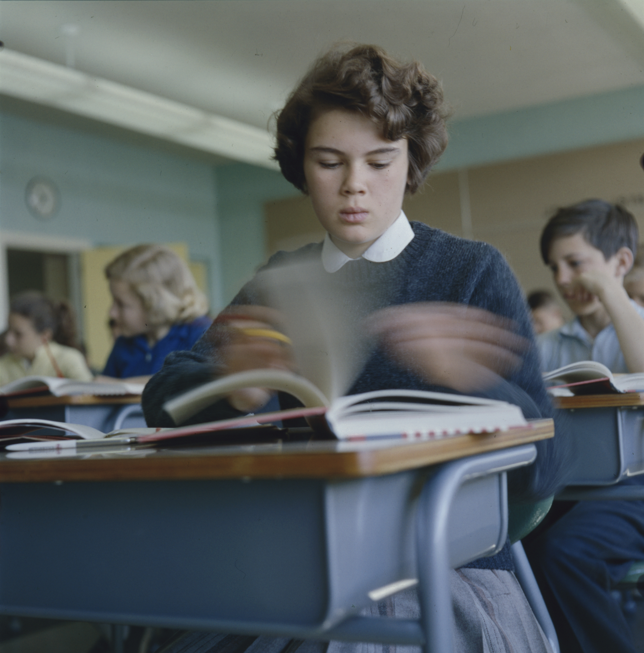 Young girl sitting at a desk flipping pages of a book in a classroom with other children.