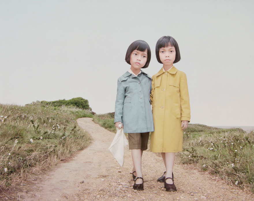 Two little girls standing on a dirt road. One is wearing a yellow coat, while the other is wearing a blue coat and holding a white scarf.