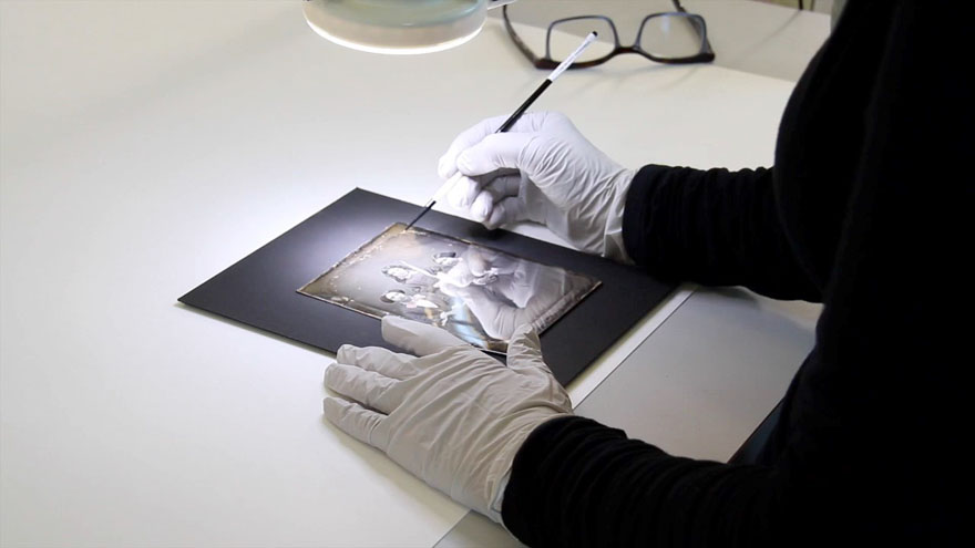 A paint brush is used under a microscope to remove mould from a photograph of three young girls. One girl is standing between two who are seated on either side.