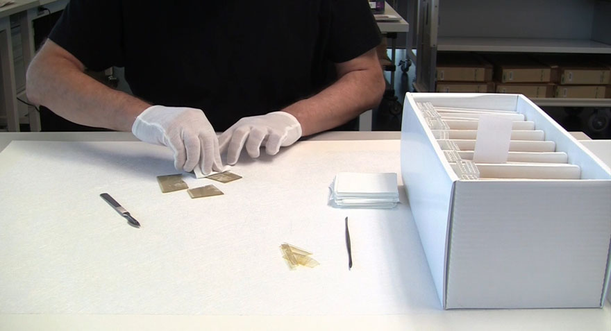 A close-up of a man wearing white gloves treating a broken glass plate negative by wiping the broken glass. Beside him is a box of glass plate negatives, in front of him are pieces of the negative.