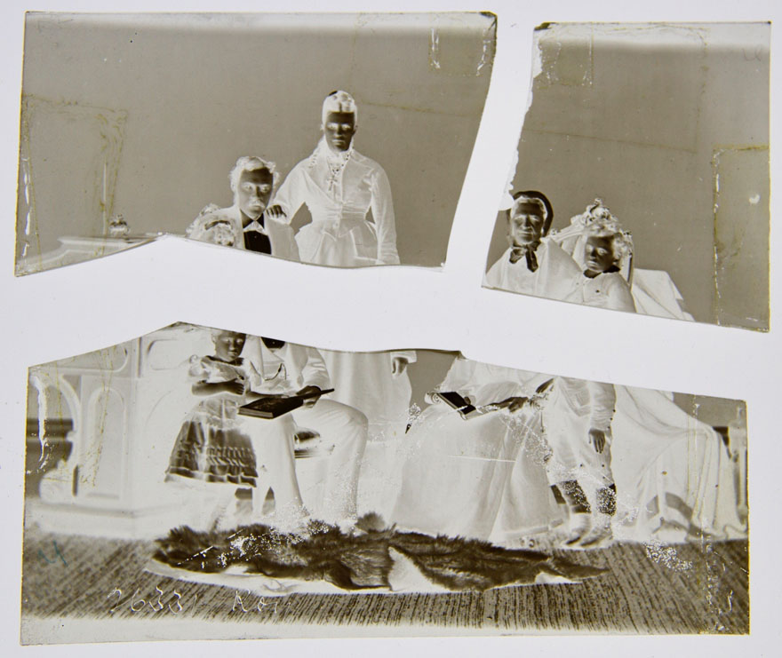 A glass plate negative broken into three pieces.  It is shown as a negative. The image is of a family portrait: a small child is standing while  reading a book with her father as he is seated, standing beside him, his wife, seated beside her an elderly woman and beside her a small boy is standing.