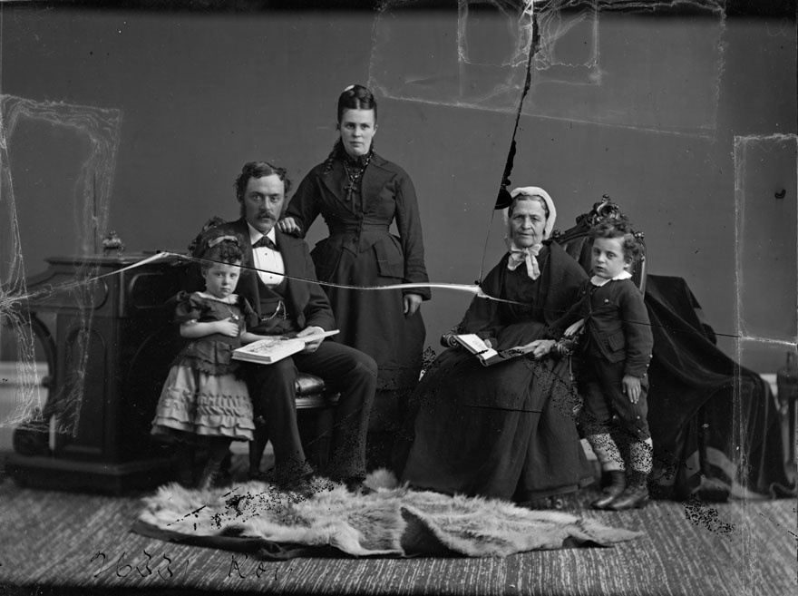 A positive of a glass plate negative. The image is of a family portrait: a small child is standing while reading a book with her father as he is seated, standing beside him, his wife, seated beside her an elderly woman and beside her a small boy is standing.