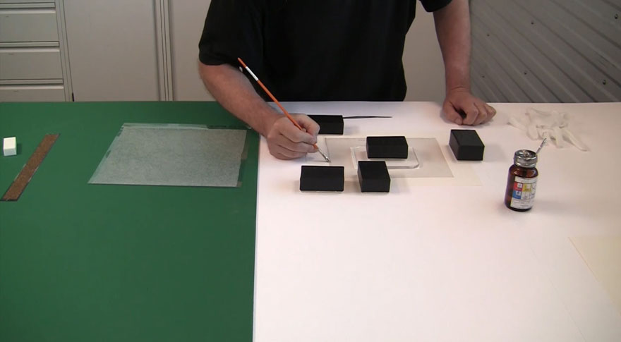 With a paint brush a man is brushing solvent onto strips of Japanese tissue onto the back of a photograph and mount. There are weights on the photograph and mount.