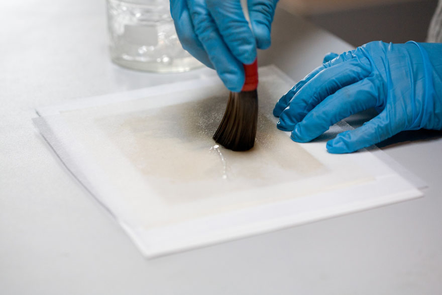 A person is brushing liquid onto a tissue that is in contact with the back of a photograph.