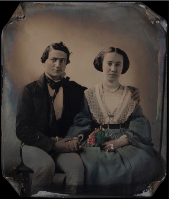 A portrait of a young couple. The gentleman has one arm around the woman and his other hand is holding hers. The woman is holding a bunch of flowers in the other. The four corners of the photographic plate are turned inward.