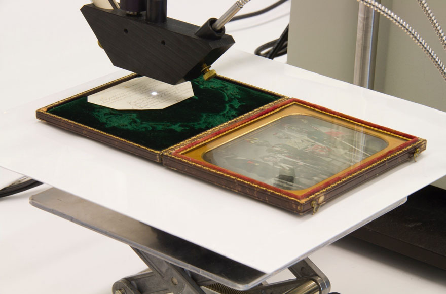 A close-up of a daguerreotype on a stand with the probe from above. A small light from the probe is piercing a handwritten inscription on the daguerreotype case.