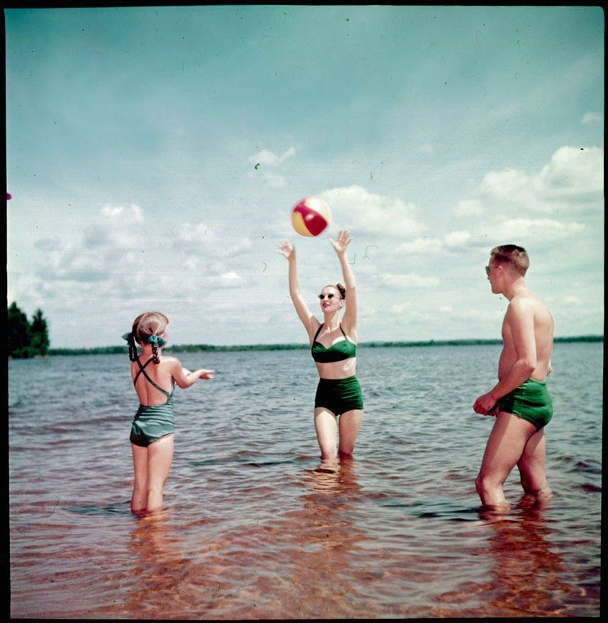 Standing in low water are three people throwing a beach ball. There is a man to the right, a woman in the centre, and a small girl to the left. All are wearing bathing suits.