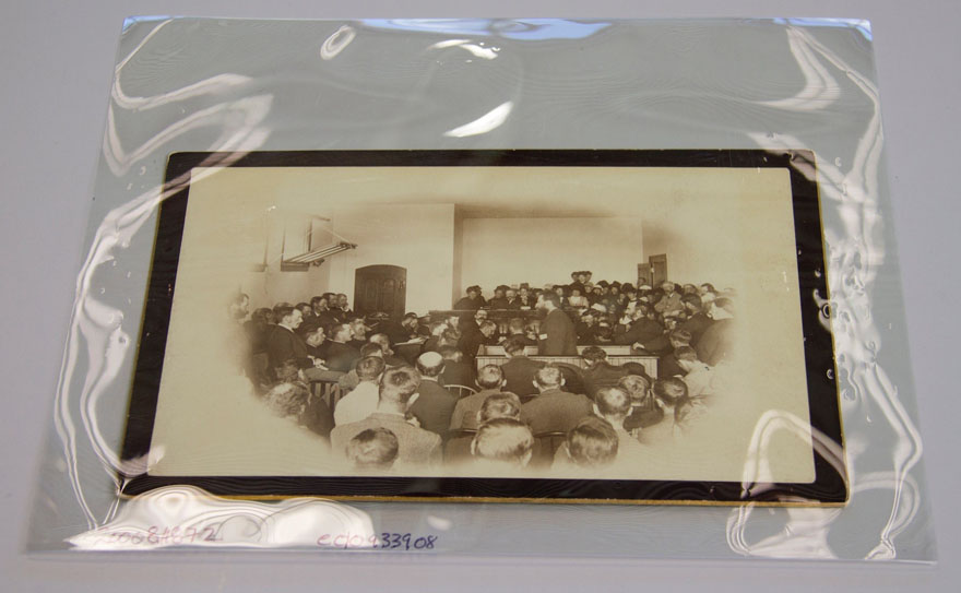 An image of a photograph housed in a plastic envelope.  In the photograph a man stands in the centre of a room. He is surrounded by other seated men, looking at him. It is a court room scene.