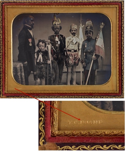 A hand coloured photograph of a man and four boys dressed in costume. The photograph is in a case with a brass mat. The bottom left edge has a stamped inscription. A close-up of a photographer's stamp 'T.C. DOANE' on a brass mat.