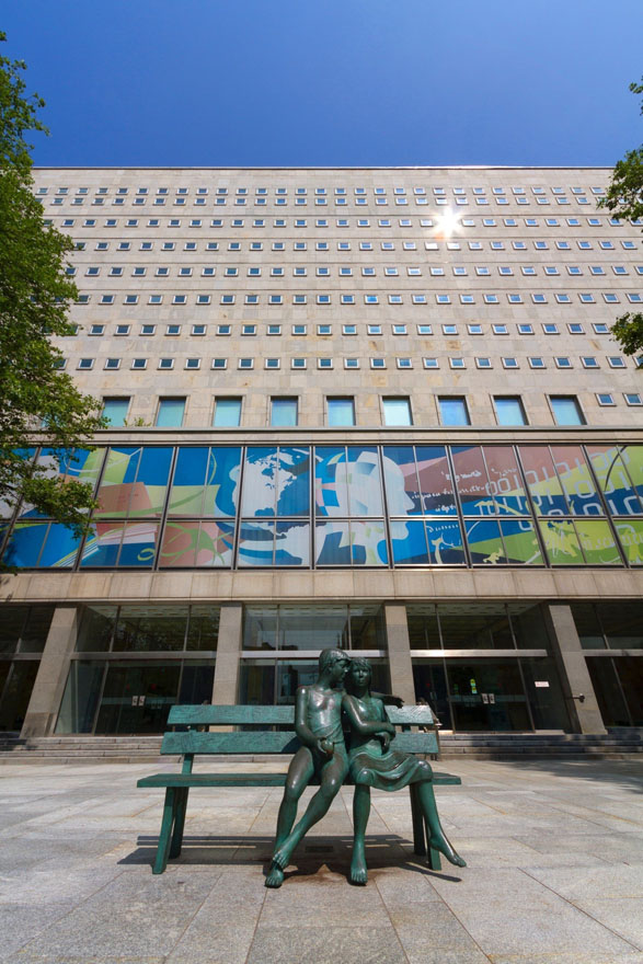 An image of the front of a large building with a bronze statue of two people sitting on a bench in the foreground.